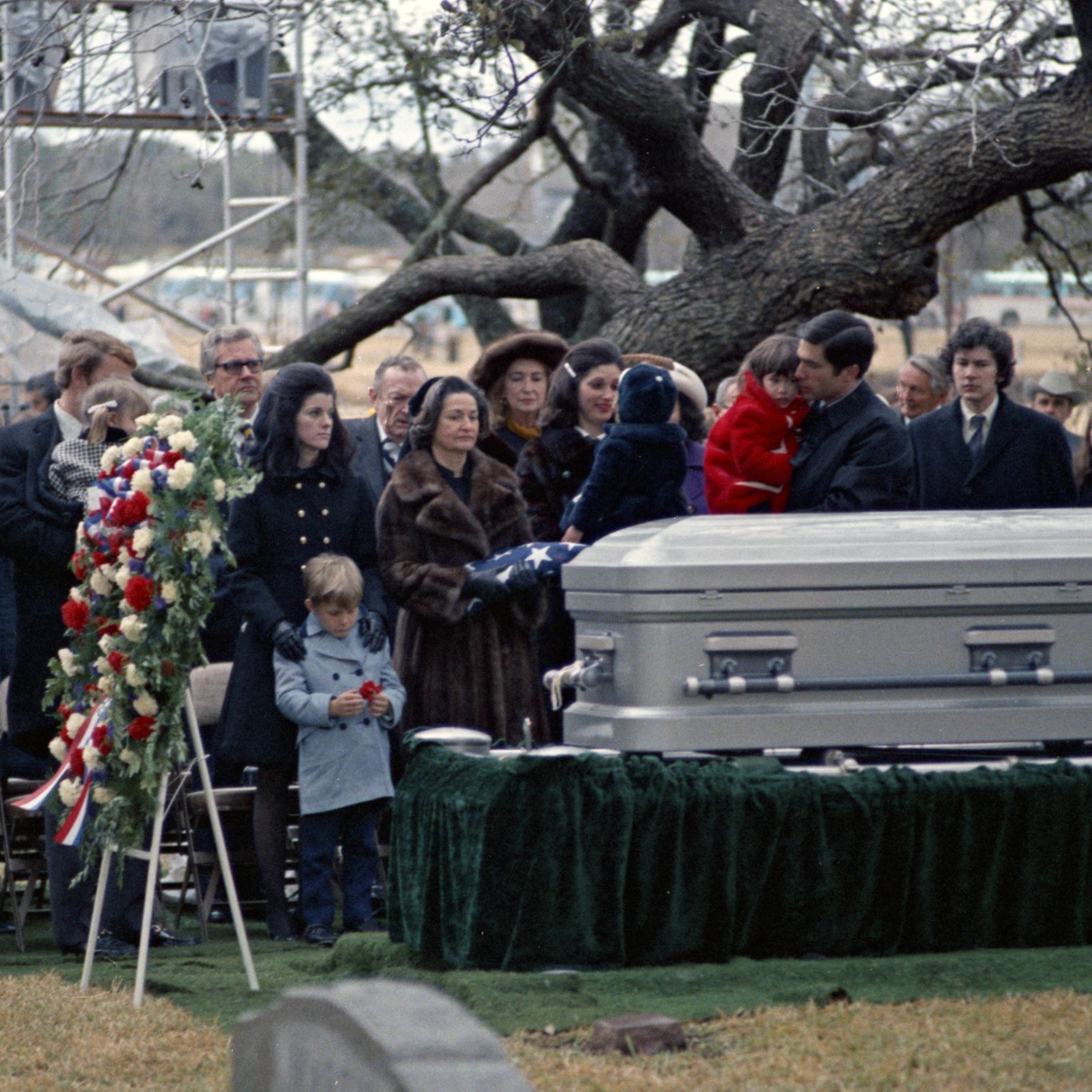 The Johnson family stand on the far side of a silver casket, a wreath and American flag in the foreground. Guests wear winter coats, except for Col. James Cross, who is in uniform.